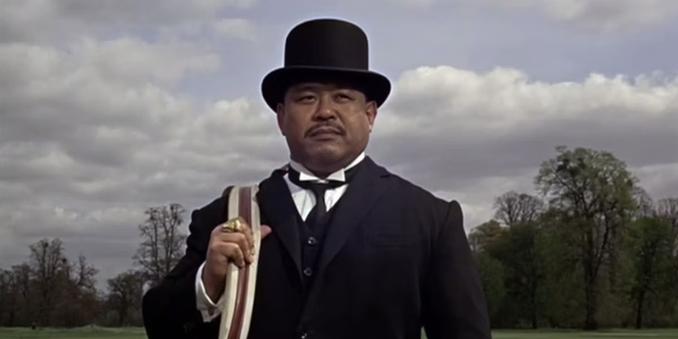 Goldfinger's handlanger Oddjob, overdressed as usual, in een smoking met een hoge hoed waarvan de rand scherp genoeg is om een tegenstander mee te onthoofden.