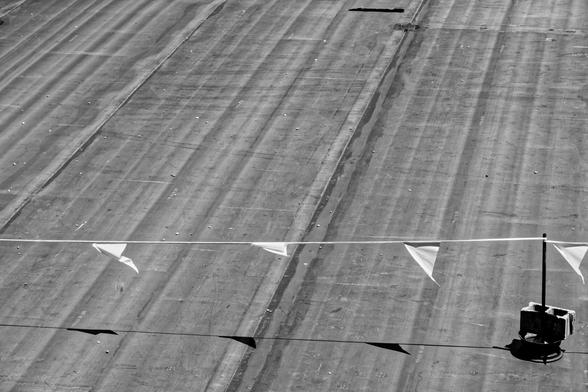A black and white photograph of a flat almost featureless roof.  On the right edge towards the foreground, a metal pole stands l held in place by a cinder block. The pole is supporting a line of triangle shaped pennants dividing the roof, traversing the frame from right to left and casting a shadow on it.