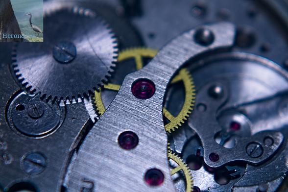 A macro perspective photograph of the inside of a pocket watch. There is a large steel gear on the upper left, with a steel arm reaching from the top left to the bottom right with three large ruby bearings embedded in it. Two brass gears can be seen underneath the bearings.