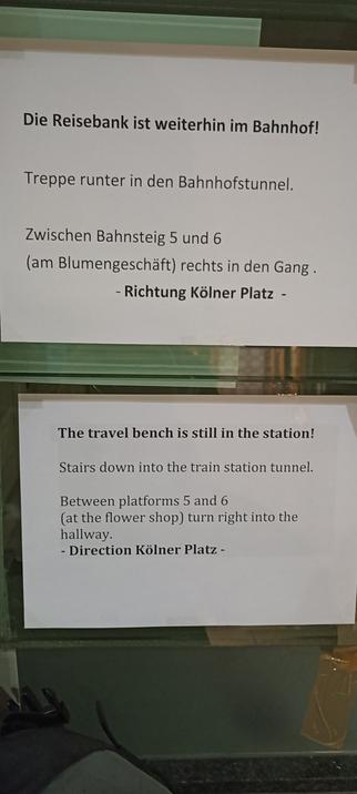 ein Aushang in dina 4 informiert darüber, dass die Reisebank an einem neuen Standort in MD Hbf zu finden ist, in der englischsprachigen Version darunter steht "travel bench"