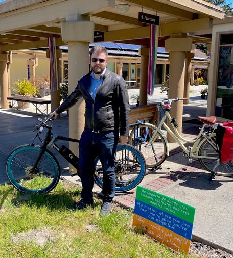 A bicycle parking rack on the courtyard is just outside the front door of the Sanctuary at First Presbyterian Church of Monterey. A man stands alongside his e-bike there, and another ebike, with panniers, is parked on the bike rack.