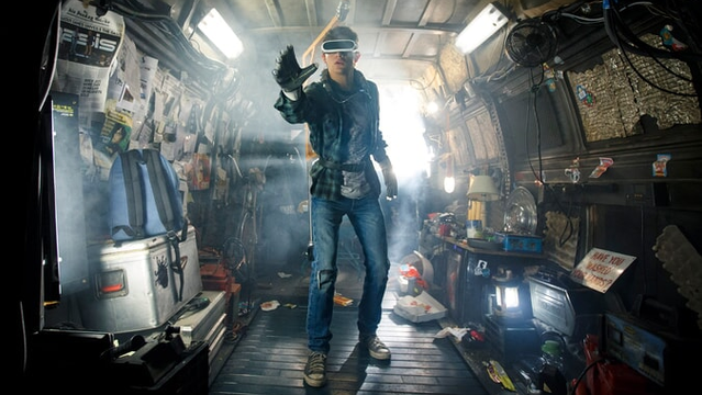 The image features a man standing in a room with a lot of clutter, possibly a garage or a storage area. He is wearing a blue shirt and a VR headset, and appears to be engaged in some activity or game. The room is filled with various objects, including a TV on the right side, a couple of chairs, and a dining table.