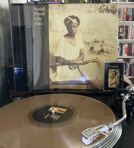 Transparent beige vinyl on a turntable with the album cover behind featuring an image of the artist seated with a axe in her lap.