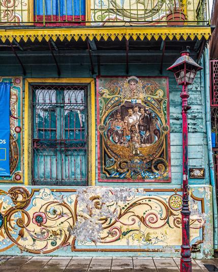 A full view of an old colorful run-down building. There is a waist-level fresco of flowers and vines, above which is a dilapidated wooden wall with a pink barred blue-green French window, next to an elaborate poster on wood topped by a woman's head over a family of five, titled ArgentinA. There's a thin red lamppost at the far right foreground - it needs paint.