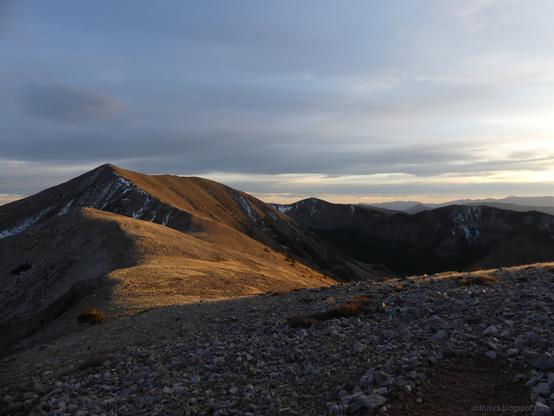 Golden late light on the peaks south of North Schell Peak in High Schells Wilderness, Humboldt-Toiyabe National Forest, Nevada.

Bare ridge line winding and rising to a point before winding a bit more. Some snow is on the surfaces on the left. The slopes drop in a steep line to green in the bottom of the valley before rising faster to another ridge, bare with a little snow on sheltered faces.