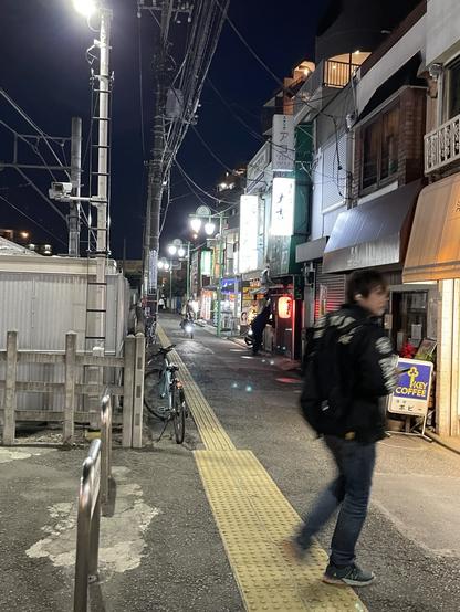 A narrow, well lighted and nearly empty street lined on one side with small shops and eating/ drinking places.