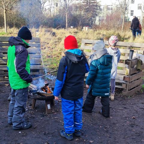 Kinder stehen um eine Feuerschale auf einem Platz mit Bänken aus Paletten.
