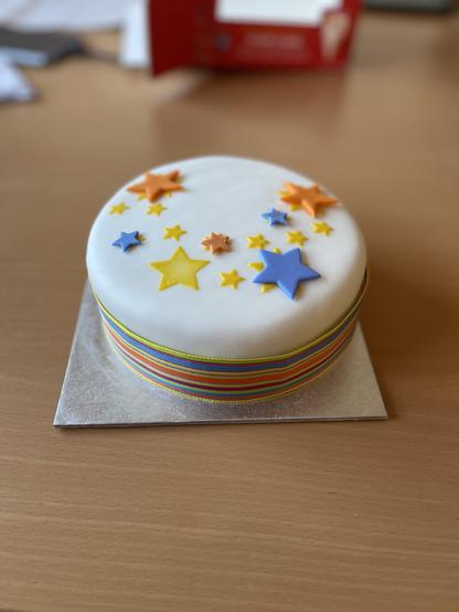 A cake decorated with white fondant and colourful stars. It is wrapped in a rainbow ribbon.