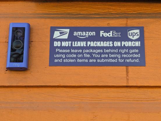 A Ring camera mounted next to a sign on an orange-painted shiplap siding. The sign has the logos for USPS, Amazon, FedEx and UPS. It reads:

DO NOT LEAVE PACKAGES ON PORCH!!!
Please leave packages behind right gate
using code on file. You are being recorded
and stolen items are submitted for refund.