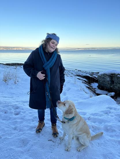 Jeg og en lysebrun labrador står på et snødekt område ved sjøen. Jeg smiler og ser ned på hunden. Har på meg en mørk vinterkåpe, blått skjerf, grått annebånd  og brune støvler. Hunden sitter og ser opp mot meg med et oppmerksomt blikk. Lyset over fjorden gir inntrykk av en sen ettermiddag eller tidlig kveld, med solen som gir en myk belysning og skaper en fredelig atmosfære. Himlen er klar og blå, og sollyset reflekteres forsiktig på vannoverflaten, noe som gir en glitrende effekt. Fjellene i det fjerne er badet i et svakt lys som fremhever deres konturer mot himmelen.