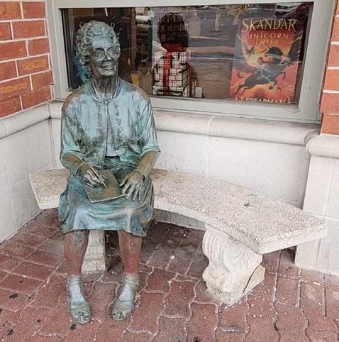 In Naperville, Illinois, USA: 
Statue/sculpture of an older lady seated on a bench, with a space for people to sit next to her. She is holding a pad of paper, with a pen poised over the paper as if writing. Her face is looking slightly up (seemingly at someone walking past or standing near), and she is smiling warmly. 

Behind the lady statue is a storefront window for a Barnes & Noble bookstore. (The bookstore is moving to another nearby town in a few weeks. I don't know what will be replacing it.)

The artist is Pamela Carpenter of Glen Ellyn, Illinois.