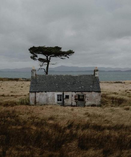 Photography. A color photograph
of an old, abandoned house in Applecross, a peninsula in Scotland. The house is gray/white and looks old and weathered. Behind it grows a green tree that looks a bit like a flat mushroom. The field around the house is covered in beige-brown grass. The sky is cloudy and a mountain range can be seen in the background. The slightly blurred gray-beige landscape with the lonely house creates a melancholy mood.
Info:
Applecross is a very old and secluded place on a peninsula that can only be reached via a very steep and winding road.