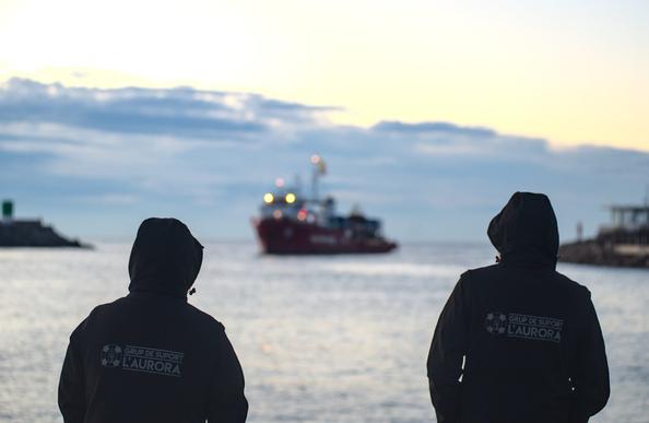 im vordergrund: zwei personen mit schwarzen jacken und der rückenaufschrift "aurora suport"
im hintergrund unscharf zwischen den beiden personen: einfahrt des rettungsschiffs seaeye4 in den hafen,