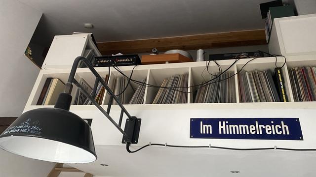 Bild einer Galerie von unten. Im Vordergrund eine schwarze Lampe, rechts daneben ein Emailliertes Straßenschild auf dem steht „Im Himmelreich“
Darüber stehen jede Menge Schallplatten in einem Regal.