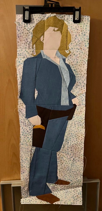 Ein Paper Piecing Bild hängt an einer Tür. Auf dem Bild ist River Song stilisiert zu sehen, sie trägt einen blauen Jeansanzug und ein braunes Halfter. Der Hintergrund sieht aus wie Lichtreflexe durch ein buntes Glas