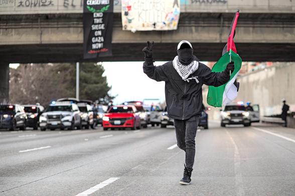 Person dressed in black wearing a keffiyeh while running with a Palestine flag in one hand, giving a Peace sign hand gesture with the other, law enforcement and banners hanging from freeway overpass seen in background. Banners Ceasefire! End the siege on Gaza now! Over eleven thousand eight hundred and thirty three children killed