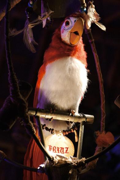 An animatronic parrot with red and white feathers on an adorned perch in a dimly lit setting.