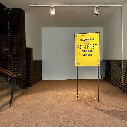 Interior of a museum, with stairs leading up on the left. The floor is covered in red clay dirt and a yellow sign reads “Willkommen Pomfret Bids You Welcome”