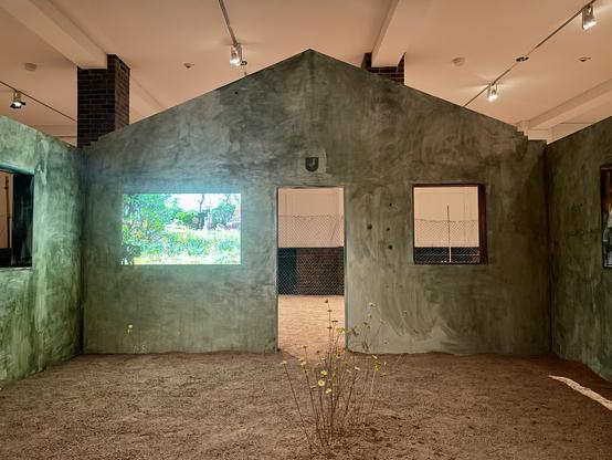 Interior of a small roofless house built inside a museum. The floor is covered in red clay dirt with a few dry flowers seeming to grow out of the ground. On one wall of the house a scene with green plants is projected.