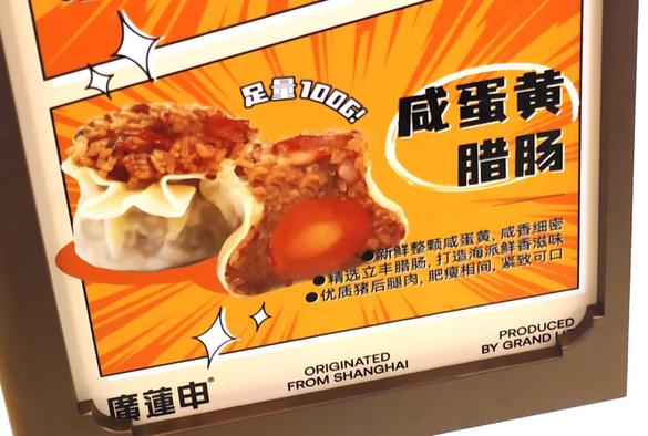 A sign advertising freshly steamed Shanghai-style shaomai sticky-rice dumpling with salted egg yolk and lap cheong sausage. The food photography shows a cross-section with a solid orange yolk and promises that each shaomai weighs at least 100g.