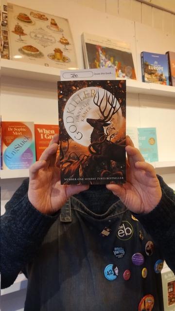 Bookseller Joe holding up a paperback copy of Hannah Kaner's Godkiller to recommend it in the Edinburgh Bookshop