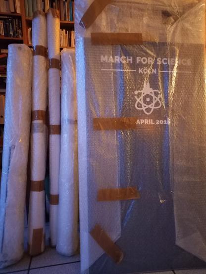 4 eingerollte Banner, in Noppenfolie verpackt, angelehnt an einen Tisch, daneben ein Plakat auf Holzrahmen, ebenfalls in Folie verpackt; das Plakat ist blau, darauf weiß der Text: MARCH FOR SCIENCE / Köln / April 2018 und ein Logo, ein Mix aus Atomsymbol und Kölner Dom