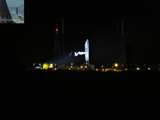 An outdoor, night time photograph of an Atlas 5 rocket, illuminated on its launch pad by harsh, white xenon lamps, venting a condensation cloud halfway up its body.
Other, yellow-orange sodium vapor lights are also visible around the rocket.
