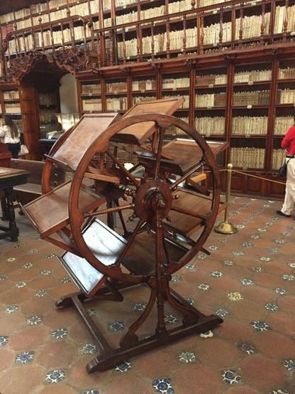 A photo of an antique wooden machine. It has a series of wooden slats between two wheels which can be rotated to keep the slats in a readable position. It's situated in an ancient old wooden library