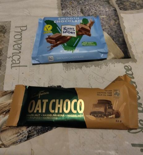 Pöydällä ylempänä Ritter Sportin vegaaninen suklaapaketti ja  Fazerin Oat Choco-vegaanisuklaapaketti.