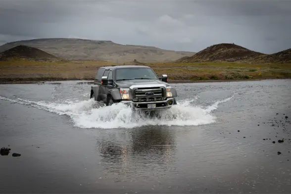 A modified Ford F350 Super Duty wades through a river in the highlands of Iceland. The vehicle creates a bow wave as it moves forward. There are little hills in the background.