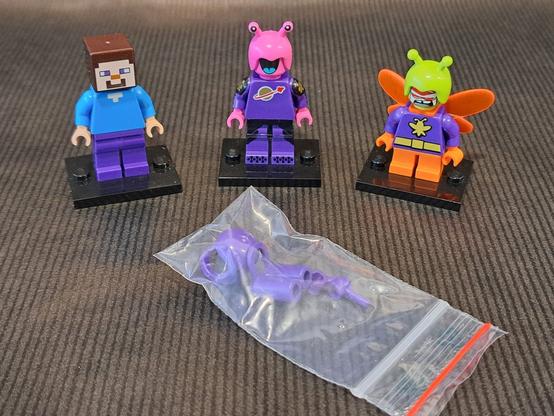 Trois figurines LEGO. À gauche, Steve de Minecraft, avec les jambes violette. Au centre, un alien avec une tête d'insecte. son torse violet est flanqué du logo Classic Space. À droite, le super vilain DC Killer Moth, avec des bras violets.
Au premier plan, un sachet plastique qui contient un casque, un réservoir et des mains violette.