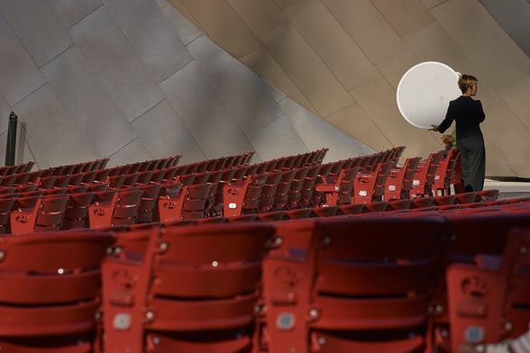 A photographer’s assistant holds a silver reflector in front of a metal wall, in the red seats of the Pritzker Pavilion in Chicago.