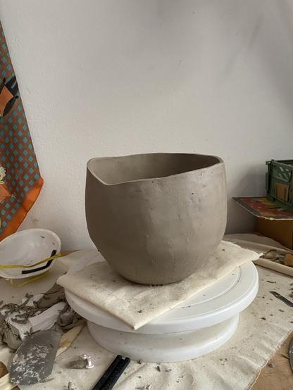 a photo of a relatively messy space with a hand-built clay vessel in between a vase and a bowl with an uneven rim, sitting on a piece of fabric and on a white turntable. The turntable is on another piece of fabric and around it is a scalpel and a blade, discarded clay trimmings and some pottery tools.
