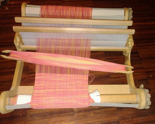 A weaving project on a small rigid heddle loom. The loom is made from light brown wood and stands dark wood floor.

The weaving is done in yellow-orange-red yarn. It produces irregular stripes and areas.