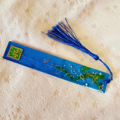 FFXIV long rectangular bookmark, Green Scholar label, cobalt tassels. Dark sapphire blue majority body with green streams, silver glitters, and fairy butterlies embedded
