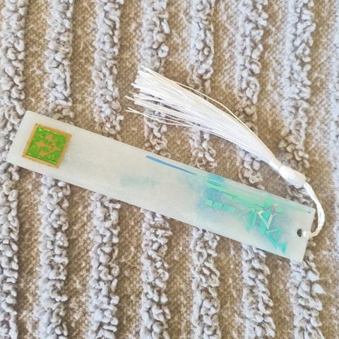 FFXIV long rectangular bookmark, Green Sage label, pearl white tassels. Blue-shimmer white majority body with aqua metalic geometric shapes, and sky blue mica hue.