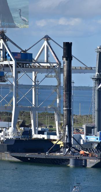 An outdoor, daylight photograph centered on a tall, cylinder that is painted white but scorched badly to black. On the base are 3 legs visible extending from the cylinder to a black deck below.
Behind the cylinder is a truss-work structure for cargo cranes and the waters of Port Canaveral beyond.