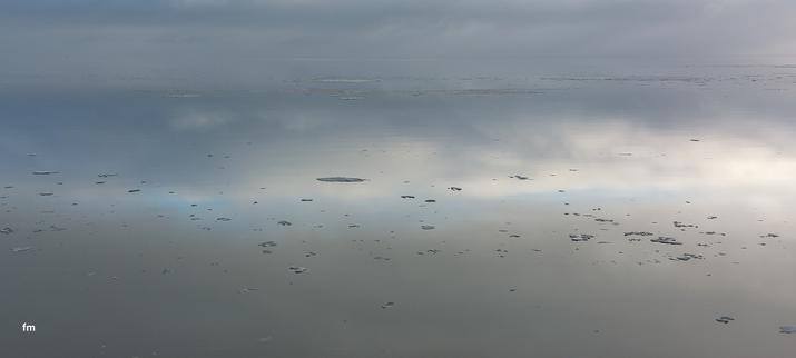 Das Bild zeigt das Wattenmeer, der Ausschnitt ist so fotografiert dass die Grenze zwischen Himmel und Wasser verschwindet. Man sieht kaum wo sich Wasser und Himmel begegnen. Die Wolken spiegeln sich im glatten Wasser, es ziehen ein paar kleinere Eisstücke vorbei.