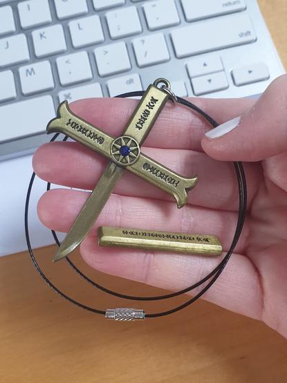 I'm holding up a merchandise version of Mihawk's tiny dagger, Kogatana. The sheath is off to show the blunt blade. The whole thing is a roughly palm sized crucifix shape, dirty gold in colour, with a bright blue gem in the center of the cross-guard.