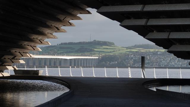 Looking over to the green hills of Fife and houses. The Tay Road Bridge is in the left portion, stretching across the River Tay. The whole view is framed by the triangular shaped walkthrough of the V&A Dundee.