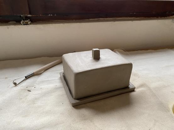 A photo of a freshly hand-built clay butter dish, resting on a off white cloth. There’s a pottery tool behind it to which I don’t know the name. It’s used for taking away excess clay. The shape of the dish is the shape of butter haha
