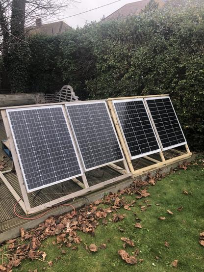 4 solar panels in wooden frames sitting on a small wooden deck in a garden. A hedge is behind them .