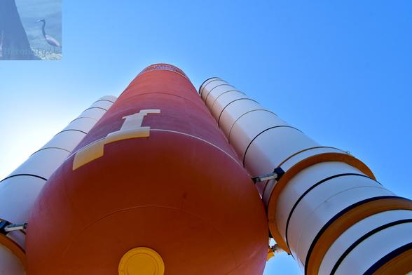 An outdoor, daylight photograph looking up to the bottom and side of an orange cylinder with two white cylinders on either side, all stretching up to the bright blue sky.