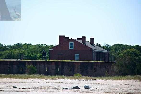An outdoor, daylight photograph of a brick building with four chimneys on its triangular roof. It has a series of black columns on the right side, and is behind a brick wall, which is behind a beach.