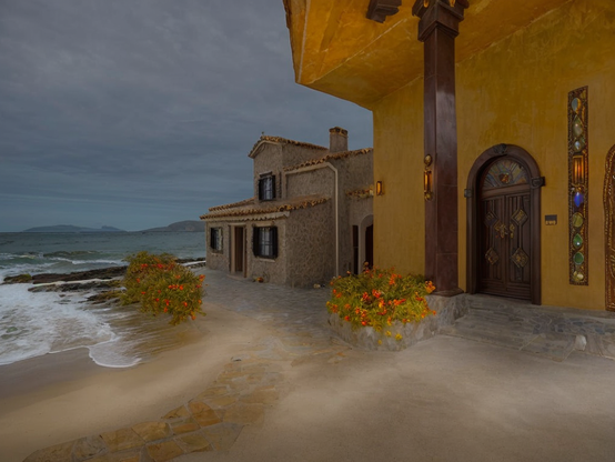 Early morning view of a beach hotel, with warm-hued walls, rustic doors, and vibrant flowers near the shore under a cloudy dawn sky.