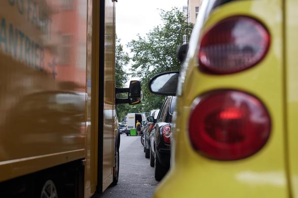 Ein Foto, das eine Straßensituation zeigt: Zu sehen ist rechts im Bild ein parkendes Auto, das offenbar auf einer Straße gemeinsam mit vielen anderen Autos in Längs-Parkrichtung parkt. Links im Bild steht auf der Fahrbahn, neben den parkenden Autos in zweiter Reihe, ein Lieferfahrzeug in gelber Farbe. Ein paar Meter vor diesem (Post-)Lieferfahrzeug steht ein weiteres Lieferauto in zweiter Reihe, bei welchem gerade etwas ausgeladen wird.