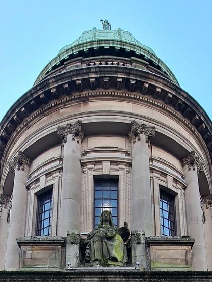 A female figure sits at the base of a rotunda above the door of a grand stone library building. At the top of the rotunda is a green copper dome topped by another female statue.