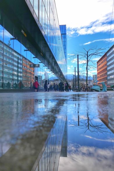 Das Bild zeigt eine belebte Straße in einer Stadt. Auf der linken Seite des Bildes befindet sich ein modernes Gebäude mit einer glatten Glasfassade, die den blauen Himmel und weiße Wolken, sowie im Hintergrund Berge und Gebäude auf der gegenüberliegenden Seite, reflektiert. Auf dem Gehweg sind mehrere Personen zu sehen. Der Gehweg ist nass und es gibt eine große Pfütze im Vordergrund des Bildes, die das moderne Gebäude und den Himmel spiegelt. Auf der rechten Seite des Bildes sind weitere städtische Gebäude sichtbar, die verschiedene Farben und Strukturen aufweisen.