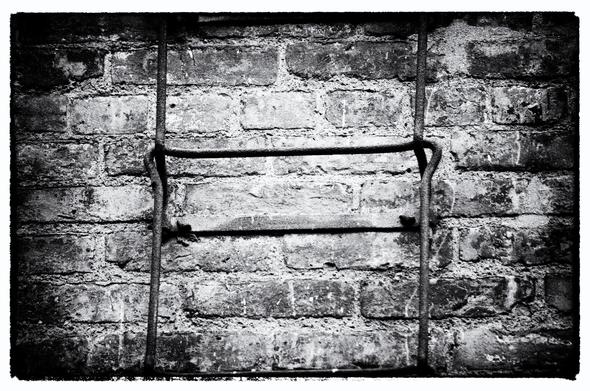 Detail of an iron fire escape against a brick wall rendered in black and white. The fire escape is made of links of iron bar, shaped roughly in a rectangle. The bottom of each link anchors into the brick of the building, bends away from the building and then runs parallel to it upward, and at the top of the link bends out a bit and wraps around the bottom of the next link.