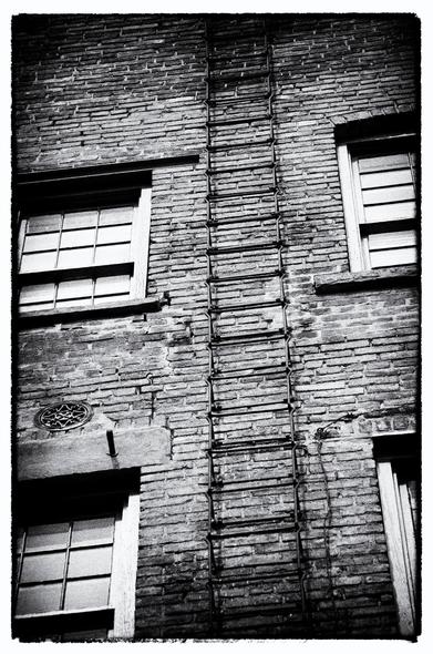 An iron fire escape against a brick wall rendered in black and white. The fire escape is made of links of iron bar, shaped roughly in a rectangle. The bottom of each link anchors into the brick of the building, bends away from the building and then runs parallel to it upward, and at the top of the link bends out a bit and wraps around the bottom of the next link. 

In this view nearly two stories of the fire escape can be seen, with old wooden mullioned windows on either side, heavy stone lintels, and decorative venting above one of the lintels.